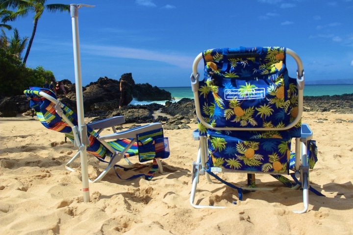 Two beach chairs on sandy shore facing the ocean with a rocky outcrop and Kihei, Wailea & Makena in the background.