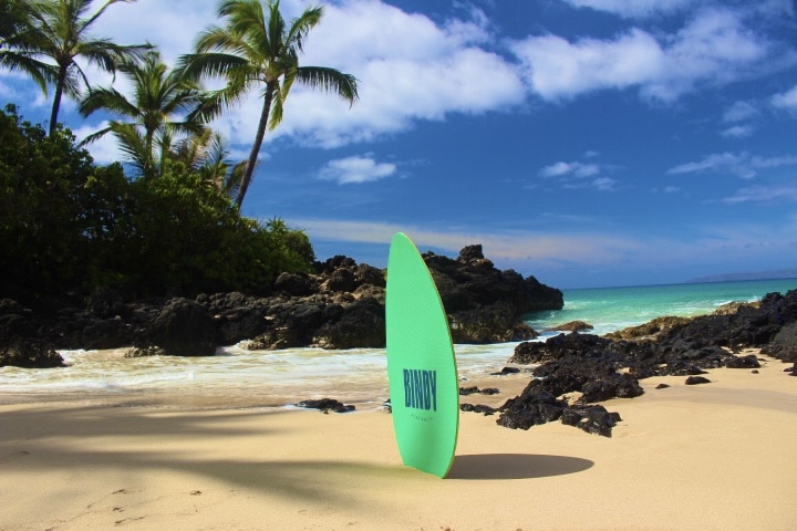 A surfboard upright on a sandy beach with palm trees and blue sky in the background, featuring GoPro Rentals Kihei, Wailea & Makena.