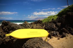 Yellow warning sign on a rocky beach with clear skies and a boat in the distance, offering Kihei, Wailea & Makena Rentals.