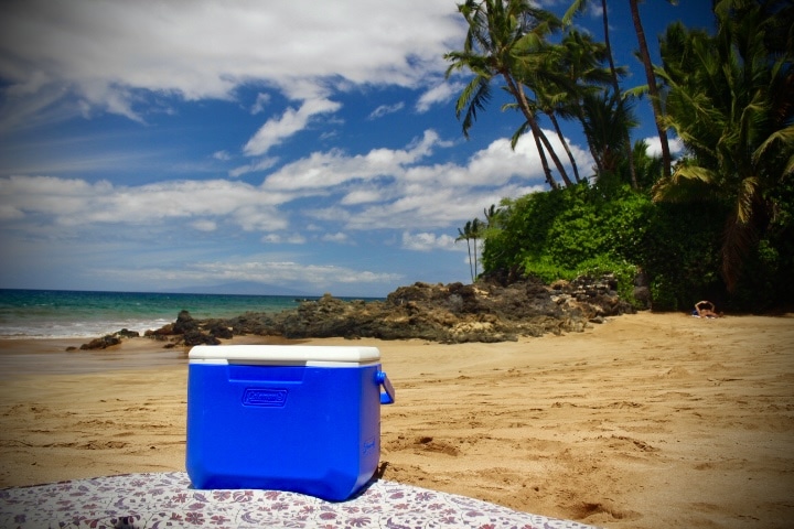 A cooler sitting on a sandy beach with tropical scenery in the background and GoPro Rentals available nearby in Kihei, Wailea & Makena.